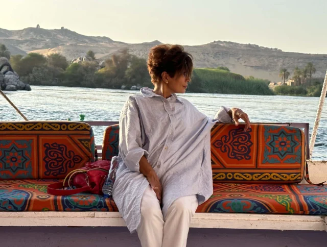 Person sitting on a colorful boat bench, looking to the side, with a river and hilly landscape in the background.