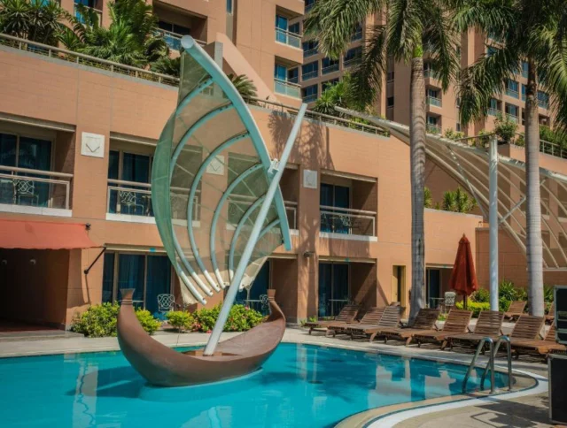 View of a hotel pool area with a sailboat-shaped sculpture, lounge chairs, and palm trees, set against a multi-story building.