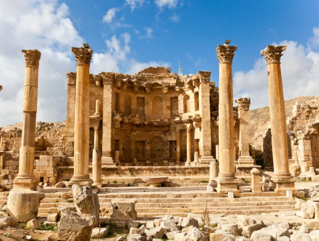 Ancient Roman ruins with tall columns and a partly intact building under a blue sky with scattered clouds.