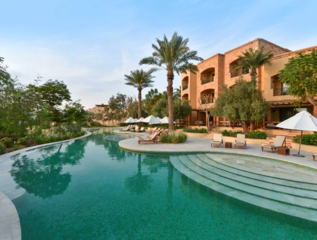 A serene outdoor resort pool curves amidst palm trees, with lounge chairs and umbrellas on a sunlit terrace beside a large villa.