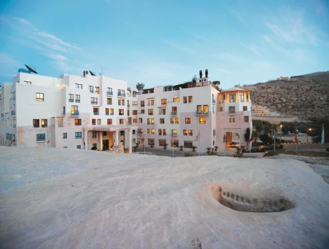A large white hotel building with multiple floors and windows, set against a blue sky, surrounded by a rocky landscape and sparse vegetation.