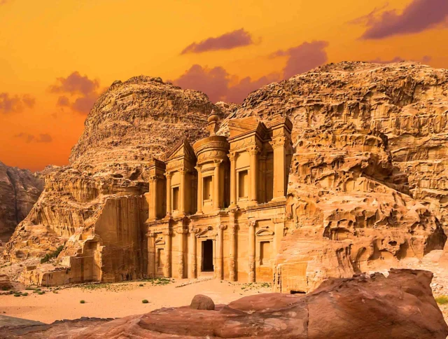 Ancient stone structure of Al-Deir (the Monastery) in Petra, Jordan, set against a backdrop of rugged cliffs under an orange sky.