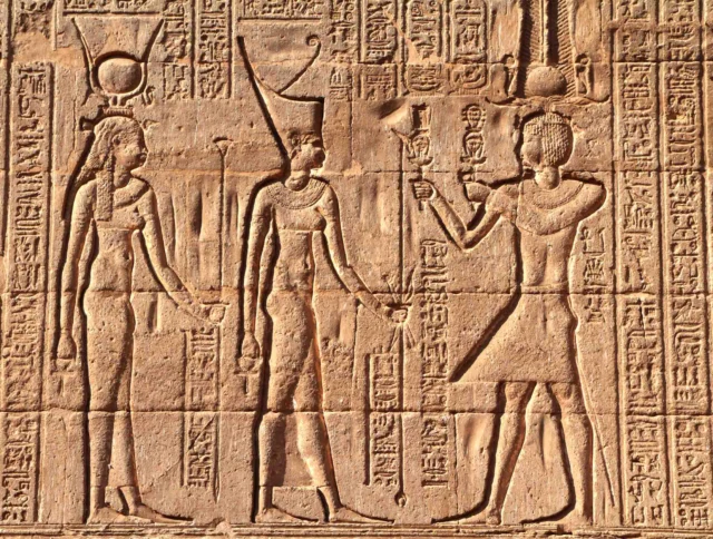 Ancient Egyptian wall carving depicting three figures in traditional attire with hieroglyphs in the background.
