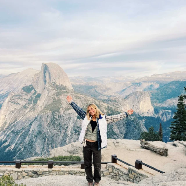 Person standing with arms raised at a mountain viewpoint, with a rock formation and trees in the background under a cloudy sky.