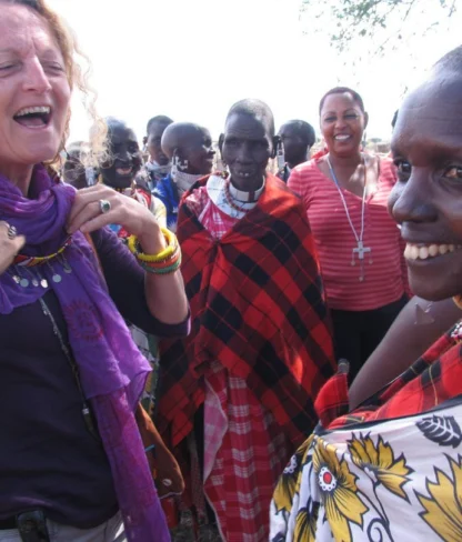 A group of people, including two women wearing colorful clothing and a man in traditional attire, are smiling and standing outdoors.