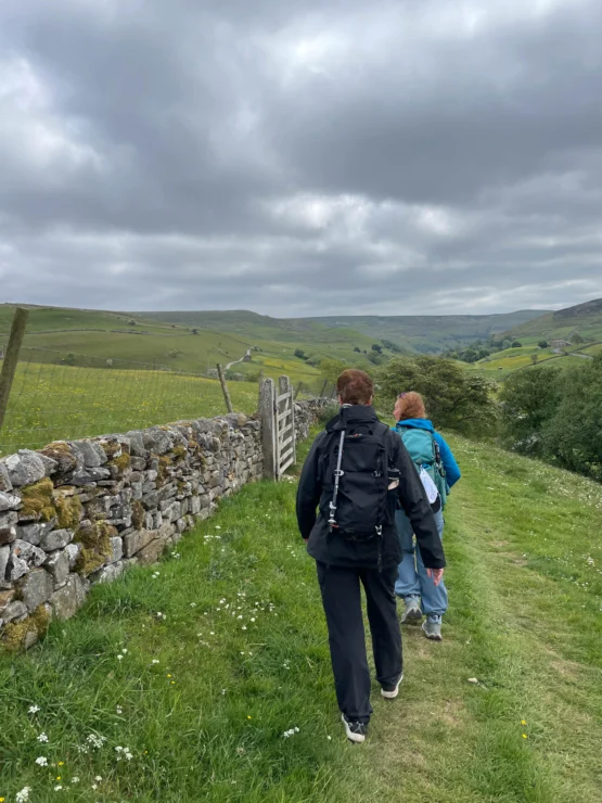 Two people walk along a grassy path beside a stone wall in a rural landscape under a cloudy sky.