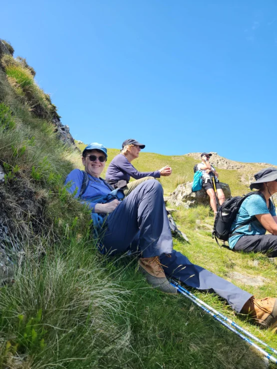 Group of hikers resting on a grassy hillside under a clear blue sky, with one person reclining and others seated nearby.