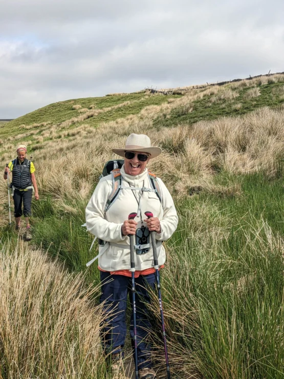 Two people hiking through grassy terrain, one wearing a hat and holding trekking poles, and another following behind.