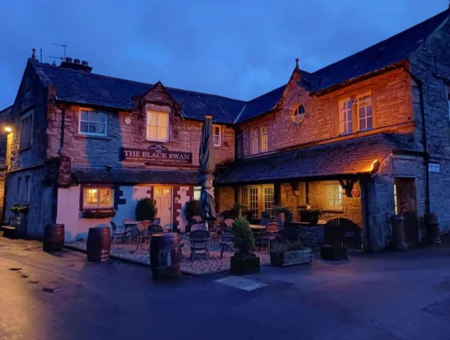 A stone building with warm lights, labeled "The Black Swan," has outdoor seating and is set against a twilight sky.