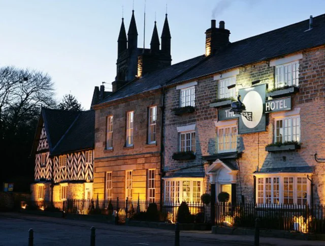 Exterior of a warmly lit hotel at dusk, with a mix of stone and timber architecture and towers in the background.