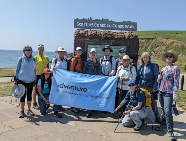 A group of hikers holding a banner at the start of Coast to Coast Walk, standing on a paved area with the sea and hills in the background.