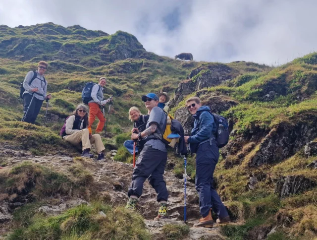 A group of people hiking up a grassy and rocky hillside, dressed in outdoor gear and carrying backpacks.