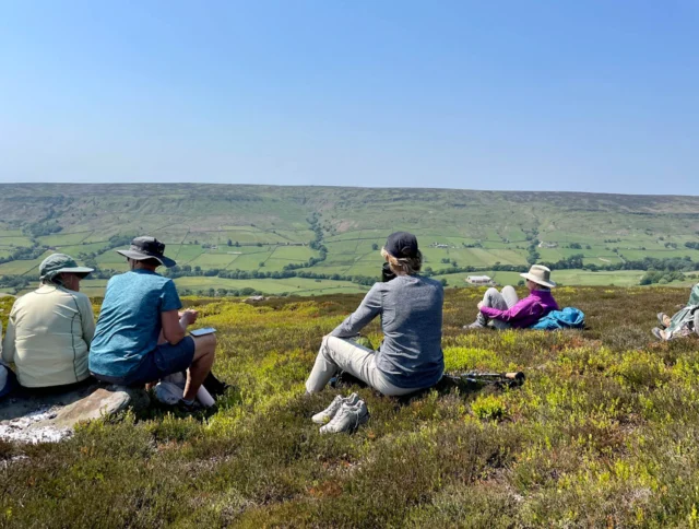 People sitting on a grassy hill, facing a vast landscape with fields and hills under a clear blue sky.