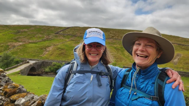 Two people in outdoor gear smiling, standing in front of a grassy landscape with a stone bridge and cloudy sky.