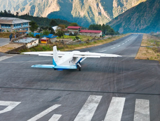 A small propeller plane on a narrow runway surrounded by mountains and village buildings in a remote area.