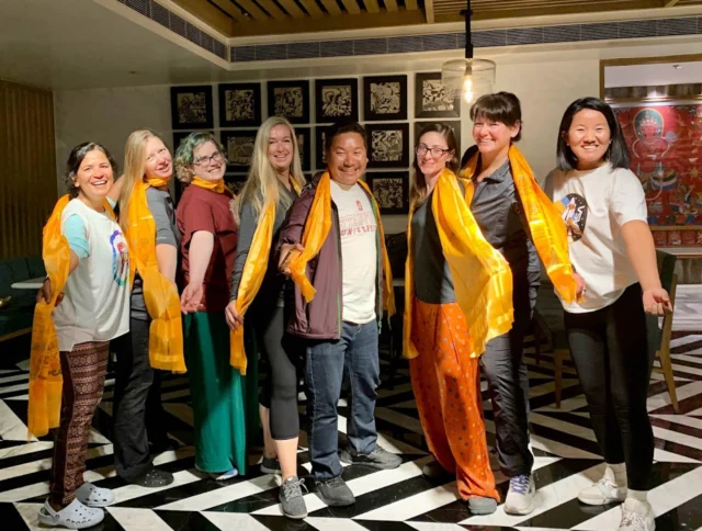 A group of eight people wearing orange scarves poses together in a room with patterned flooring and artwork on the walls.
