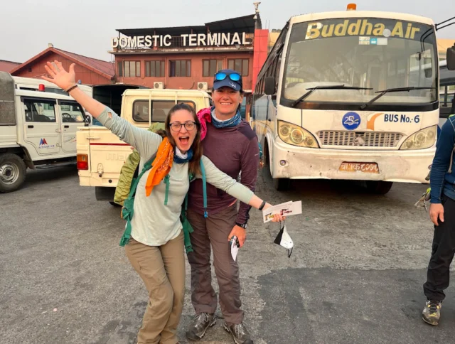Two people smiling and posing in front of buses at a domestic terminal. One person has arms raised, and they both wear hiking attire and sunglasses.