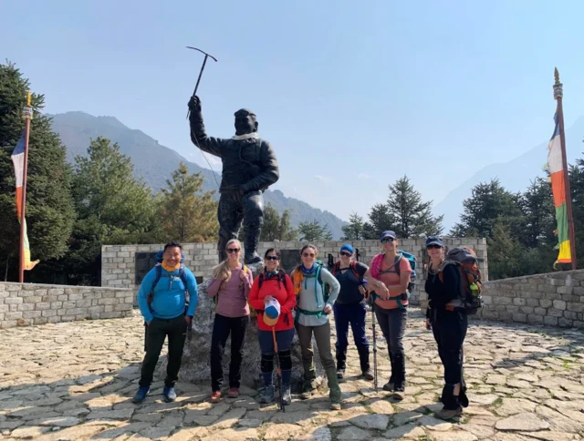 A group of people posing in front of a mountaineer statue holding an ice axe, surrounded by flags and trees, with mountains in the background.