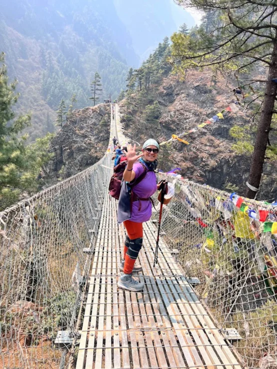 Person waving while standing on a narrow suspension bridge adorned with colorful flags, surrounded by mountainous landscape.