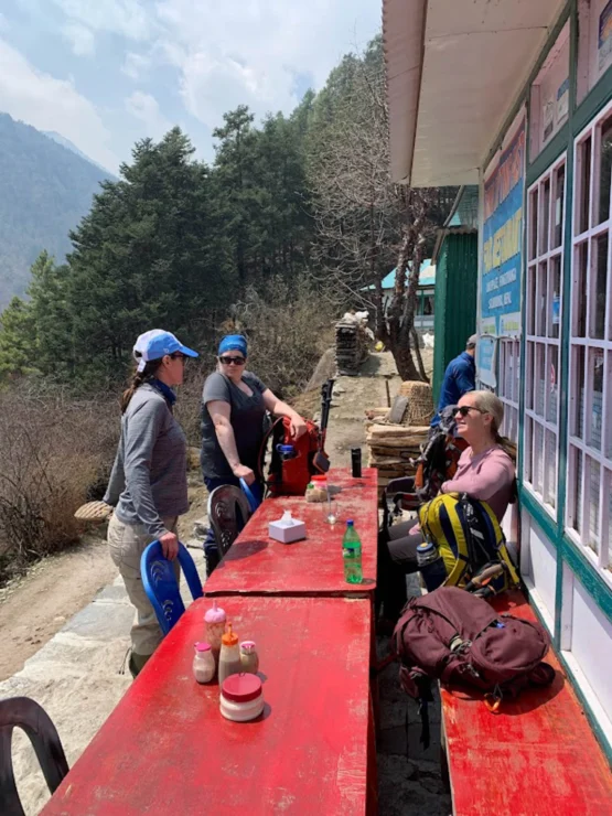 People with backpacks resting at a red table outside a building in a mountainous area.
