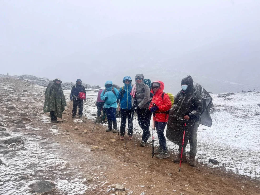 A group of people in winter clothing stands on a snowy trail with trekking poles, surrounded by misty mountains.