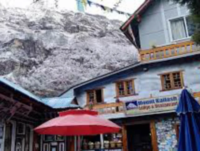 A lodge and restaurant with a red umbrella sits in front of a snow-dusted mountain under a clear sky. Tibetan prayer flags hang above.