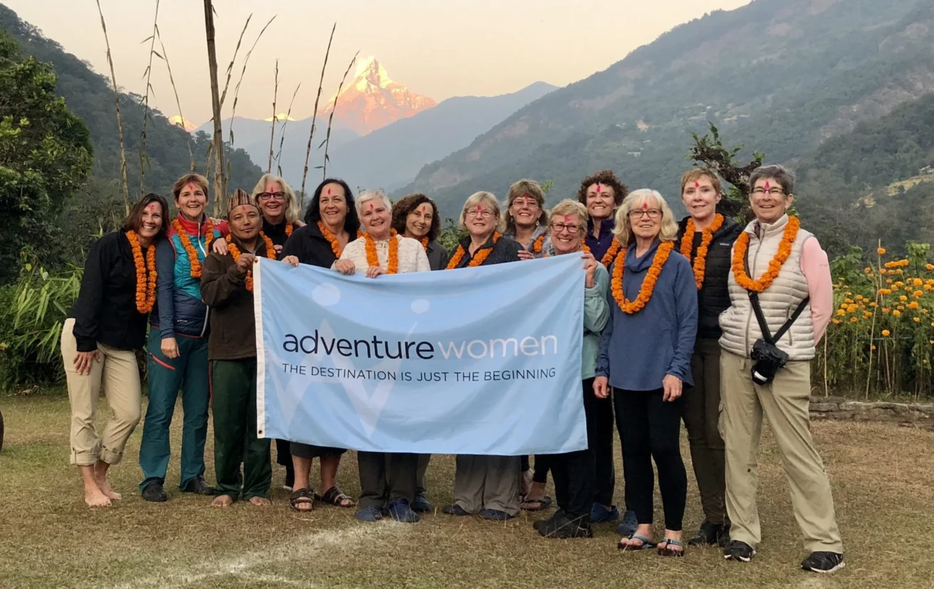 A group of women wearing orange marigold garlands hold an "Adventure Women" banner outdoors with a mountain in the background.