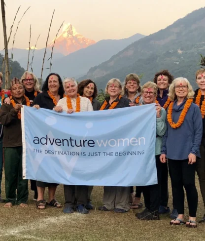 A group of women wearing orange marigold garlands hold an "Adventure Women" banner outdoors with a mountain in the background.