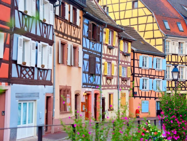 Colorful half-timbered houses with various pastel hues line a street. Bright flowers are in the foreground, adding to the vibrant scene.