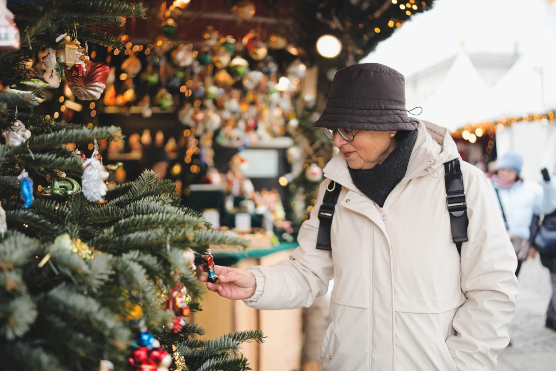 An elderly person in a beige coat and black hat examines ornaments on a decorated Christmas tree at an outdoor market.