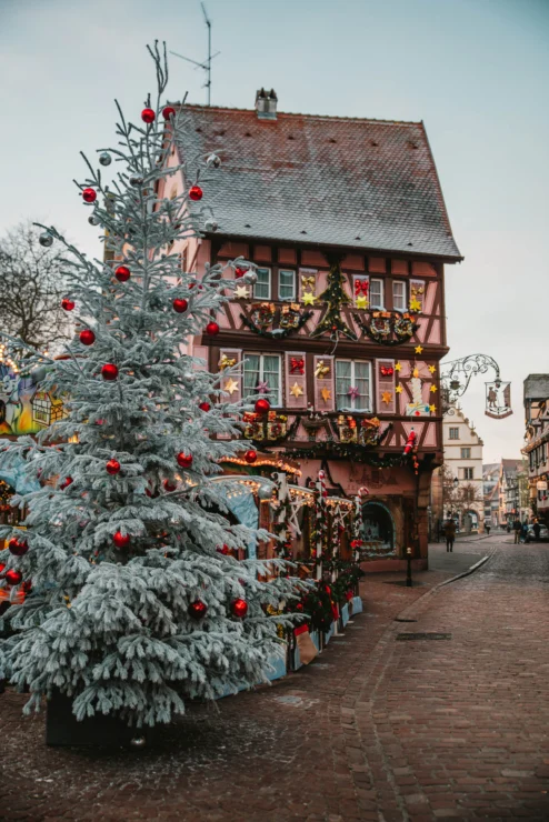 A decorated Christmas tree stands in front of a charming, timber-framed building adorned with festive garlands and ornaments on a cobblestone street.