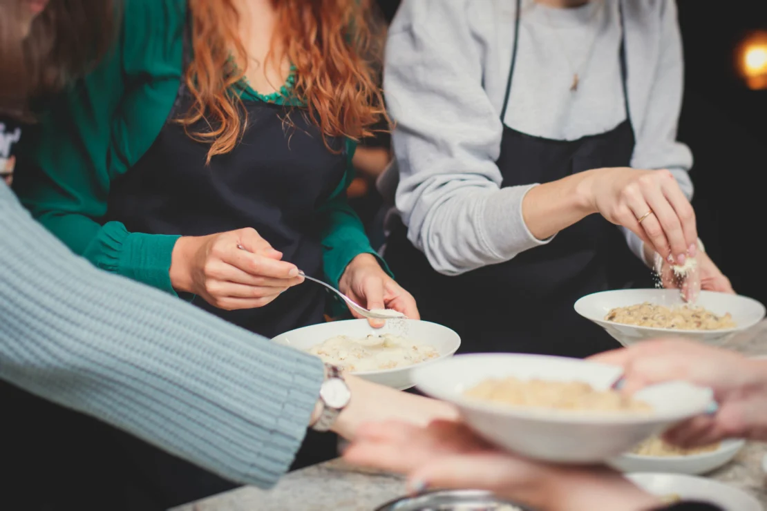People serving food into bowls, wearing aprons and using spoons, in a collaborative cooking setting.