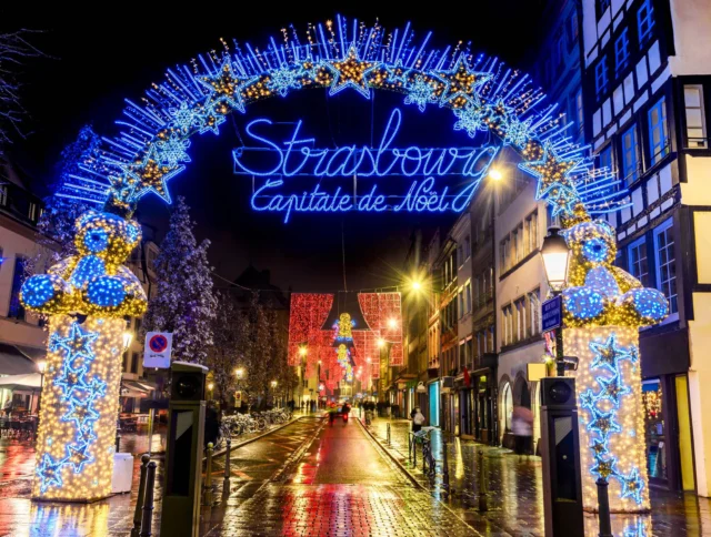 Illuminated archway with "Strasbourg Capitale de Noël" in blue neon lights, festive decorations, and lights adorn a wet street lined with buildings at night.
