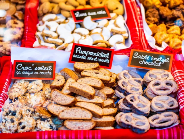 An assortment of cookies displayed on trays, including pumpernickel, croq soleil, and sablé pretzel chocolat, with labels on a red cloth.