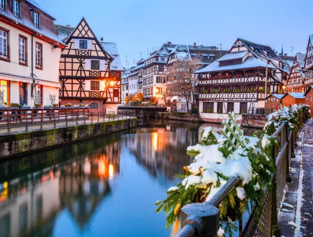 A picturesque winter scene of a quaint town with half-timbered houses by a canal, dusted with snow. A bridge and decorated railings are visible under a clear, twilight sky.