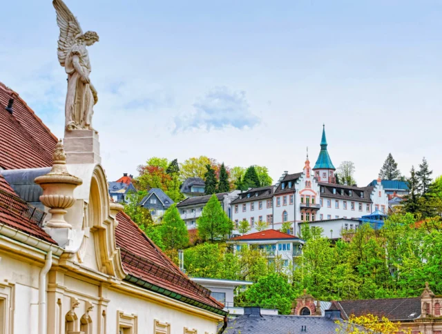 A scenic view showcases a statue on a building's rooftop in the foreground, with a hillside town featuring trees and a church spire in the background.