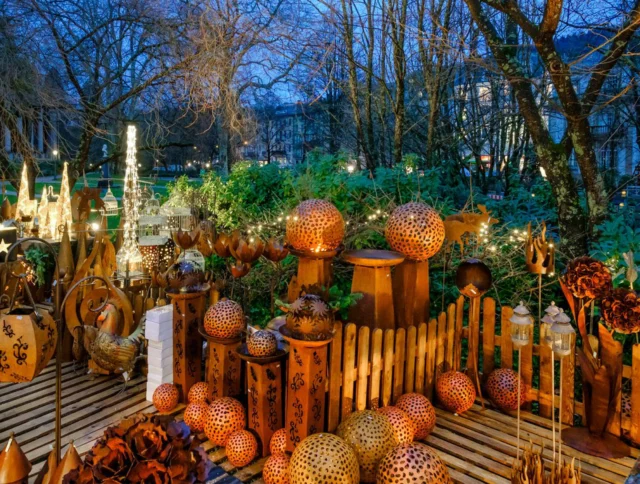 Outdoor market display with lanterns, decorative metal spheres, and wooden items on a fenced platform, set against a backdrop of trees and evening lights.