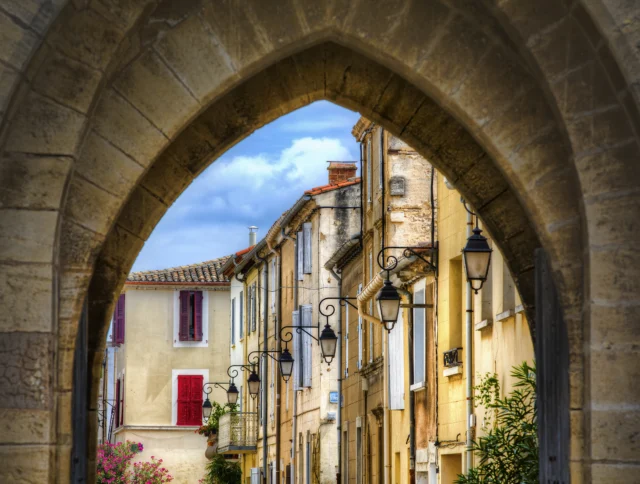Stone archway framing a narrow street with rustic buildings, hanging lanterns, and colorful shutters under a cloudy sky.