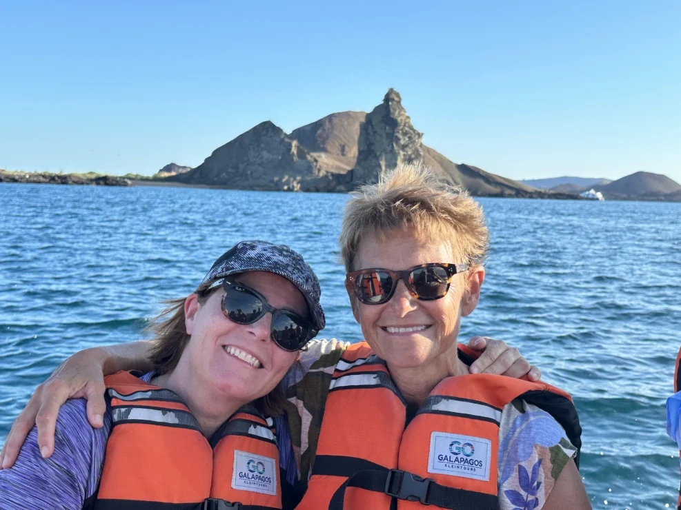 Two people wearing life vests sit on a boat in front of a rocky island formation in the Galapagos, with clear blue water and sky in the background.
