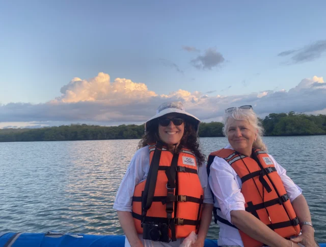 Two women wearing orange life jackets sit on a boat against a backdrop of calm water and clouds. One holds a camera.