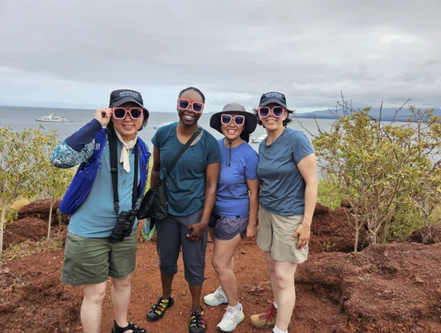 Four people in casual outdoor attire and sunglasses pose together on a rocky path near the ocean. A boat is visible in the distance under a cloudy sky.