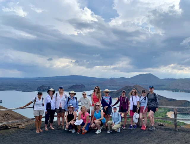A group of people posing on a hilltop with hats and casual attire. A scenic view of a bay and mountains is in the background under a cloudy sky.