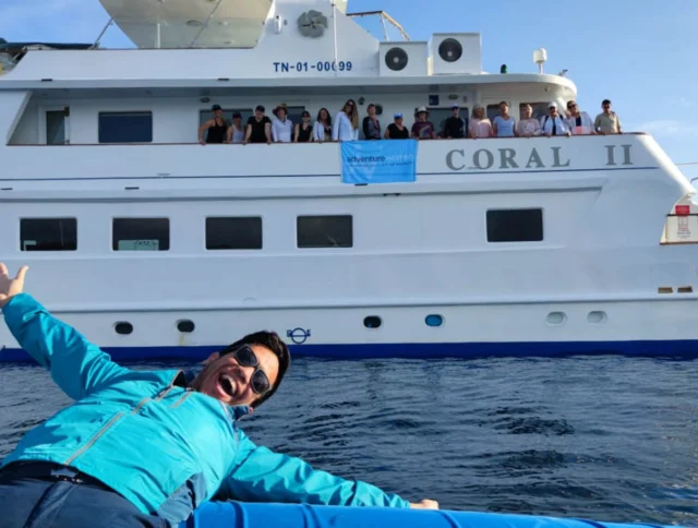 A person in a blue jacket poses playfully in front of a large white boat named "Coral II," with people standing on the deck.