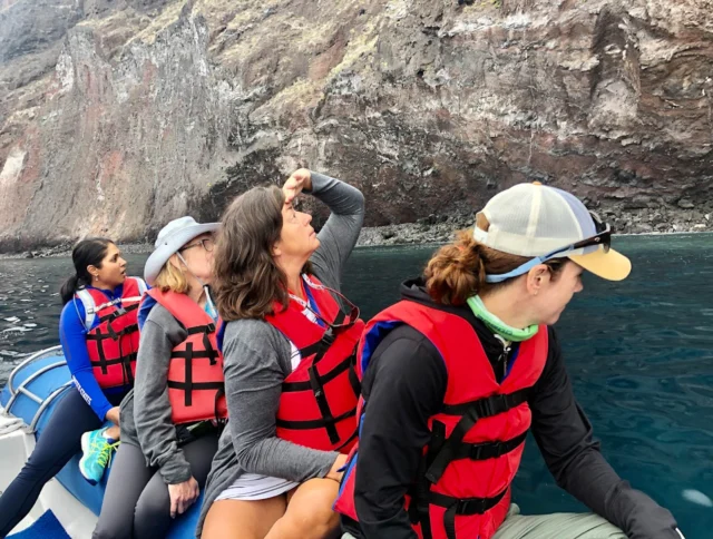 Four people wearing red life jackets sit on a boat near a rocky cliff, with one person pointing towards the sky.
