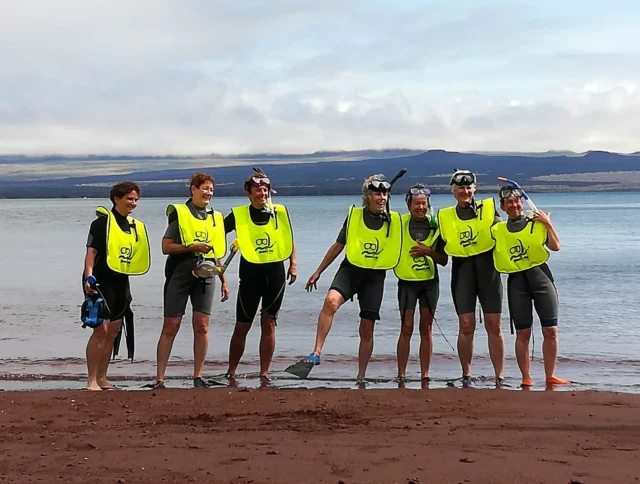 Seven people in wetsuits and yellow vests pose on a beach with snorkeling gear, against a backdrop of water and distant hills.