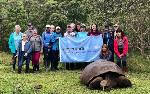 A group of people stand outdoors holding a banner that reads "adventure women" near a large tortoise on the grass.