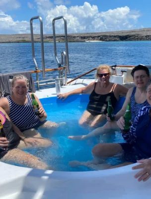 Five people relaxing in a small pool on a boat, holding drinks, with a life preserver in the background and a coastal landscape visible.