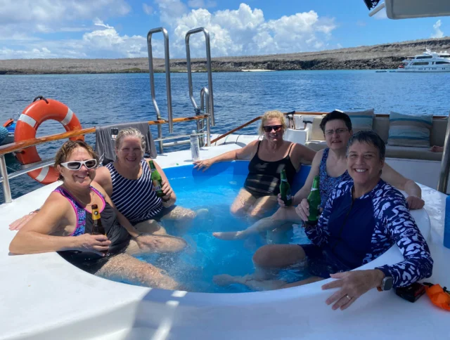 Five people relaxing in a small pool on a boat, holding drinks, with a life preserver in the background and a coastal landscape visible.