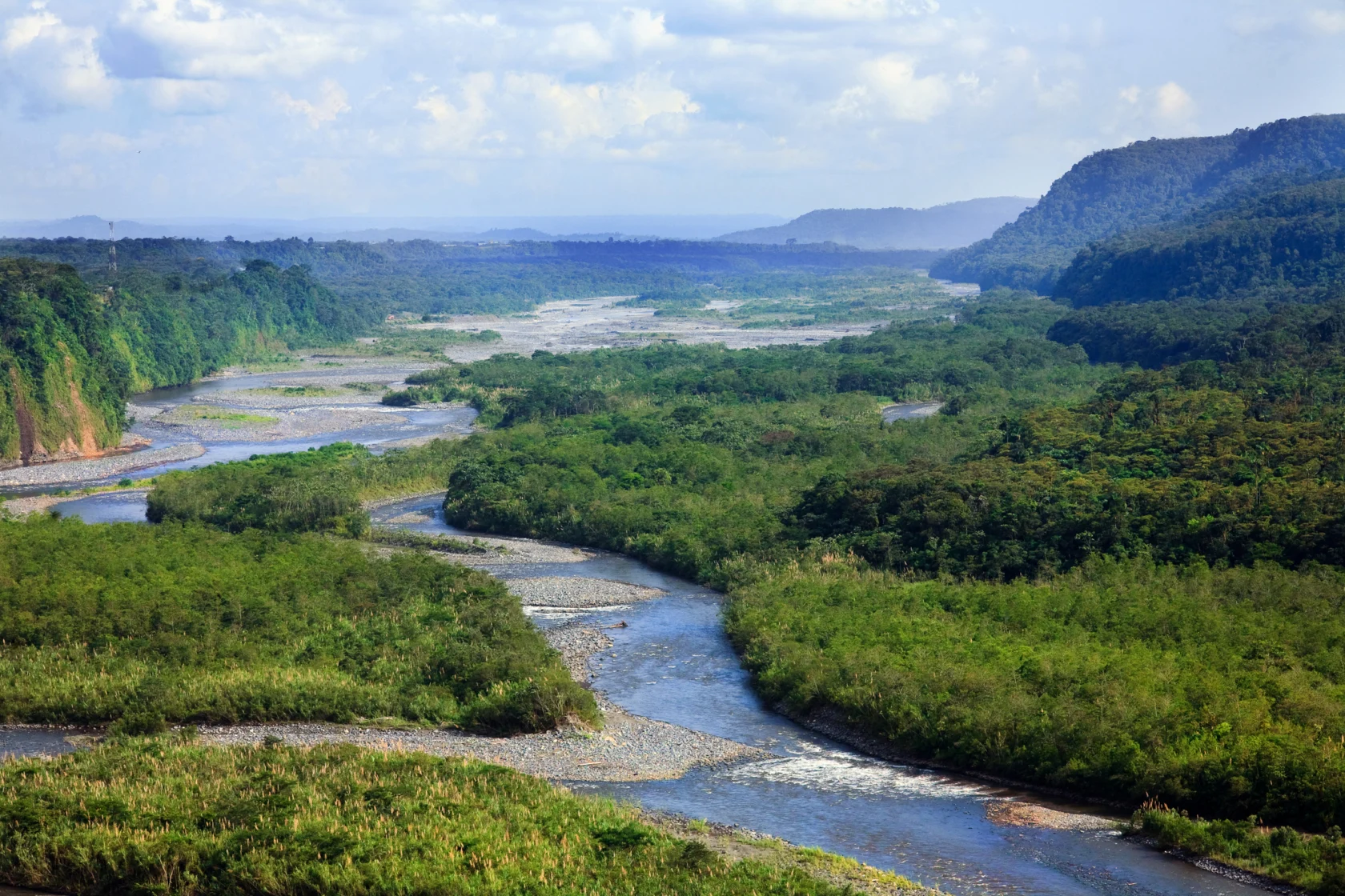 Aerial view of a winding river flowing through a lush green valley with hills in the background under a partly cloudy sky.