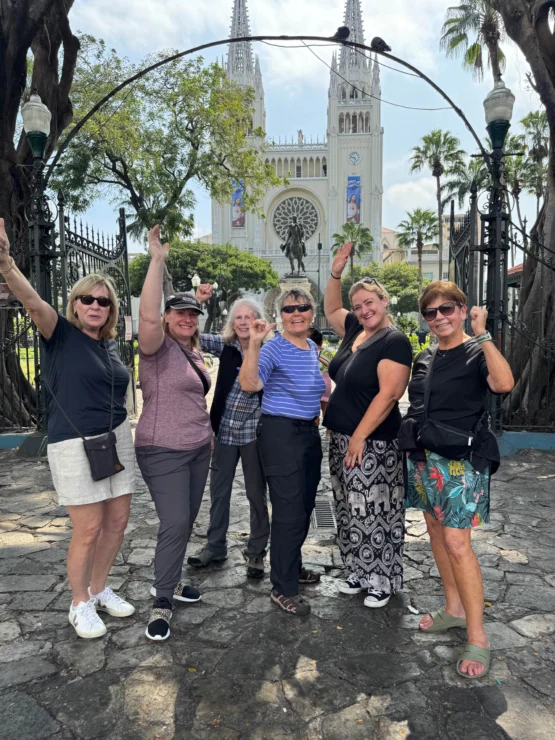 A group of six women pose happily in front of a cathedral with gothic architecture, surrounded by trees and an ornate iron gate.